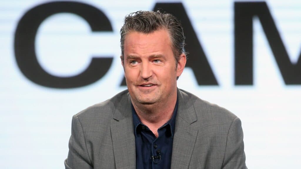 Matthew Perry durante una presentación en un evento, vestido con traje gris y camisa oscura, en el contexto de su carrera en televisión.