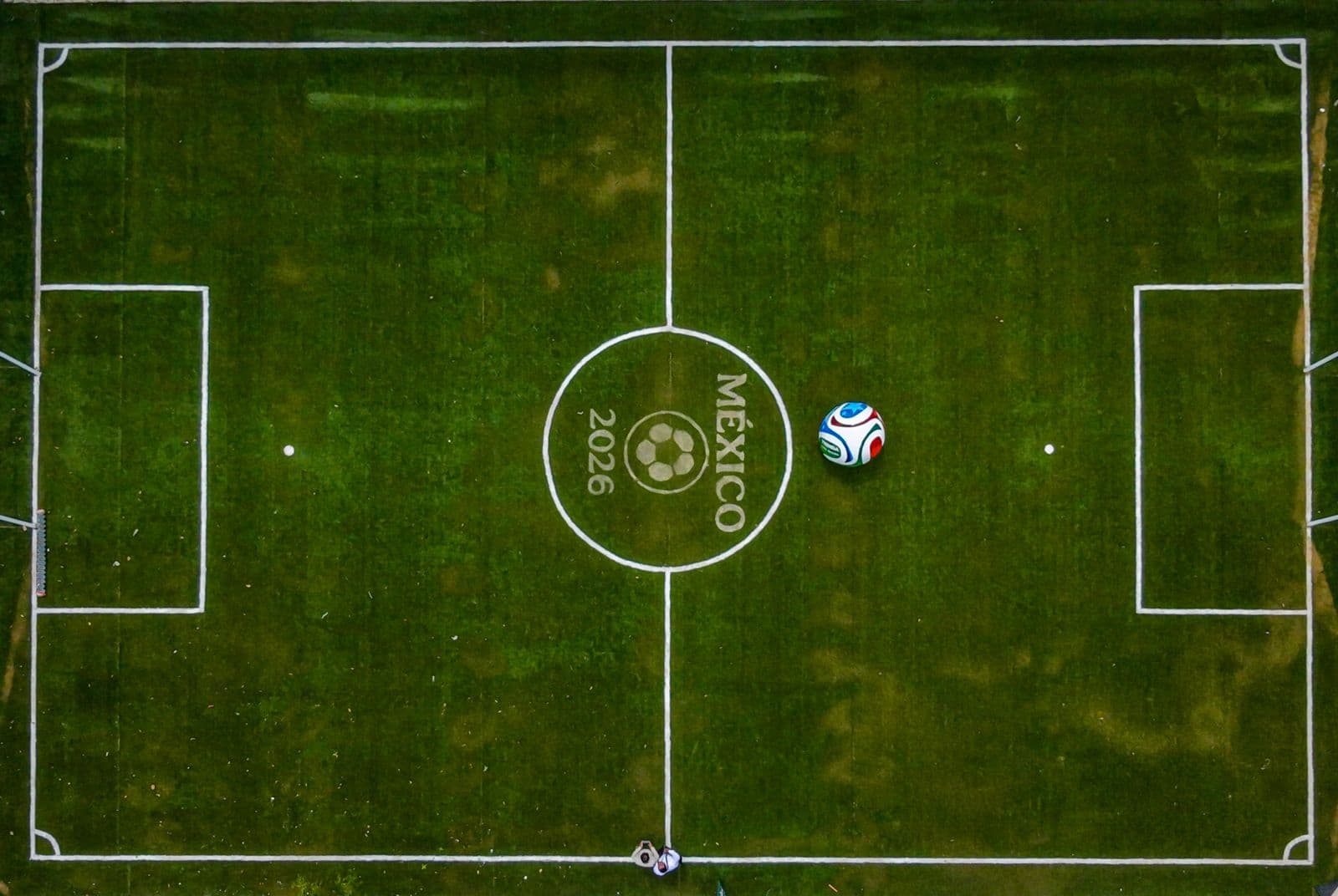 Campo de fútbol con el logo de México 2026, un balón en el centro y líneas de juego bien definidas.