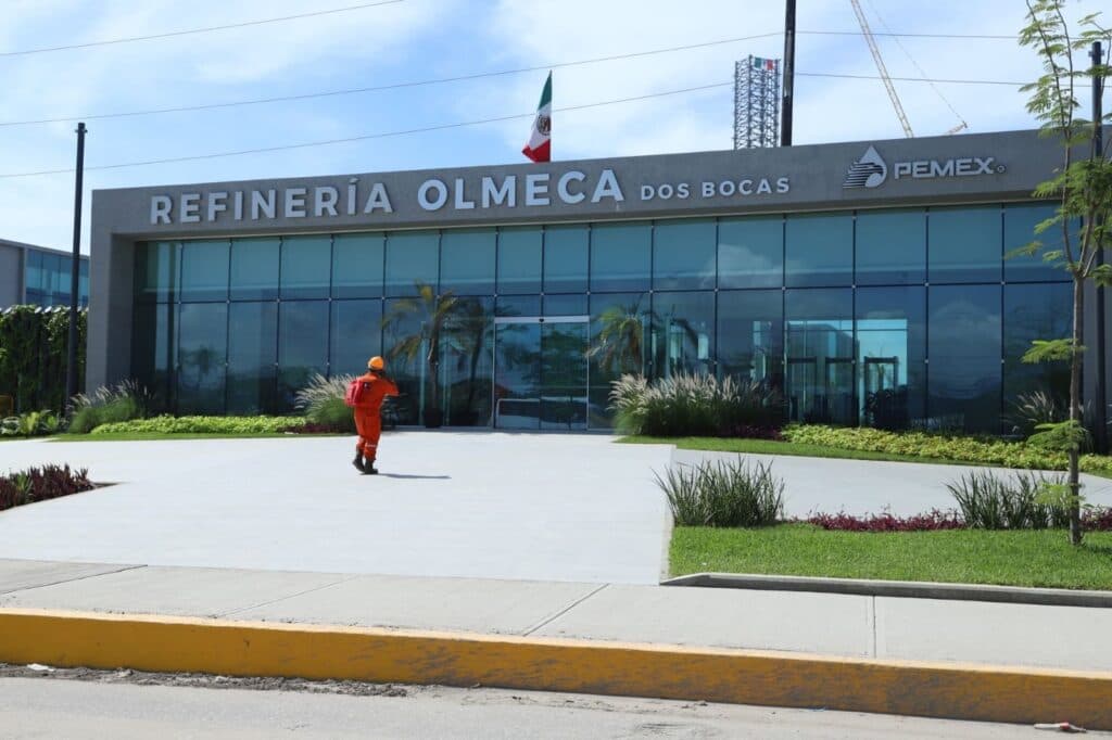 Trabajador ingresando a la Refinería Olmeca en Dos Bocas, Tabasco, con bandera mexicana ondeando y edificio moderno de Pemex al fondo.