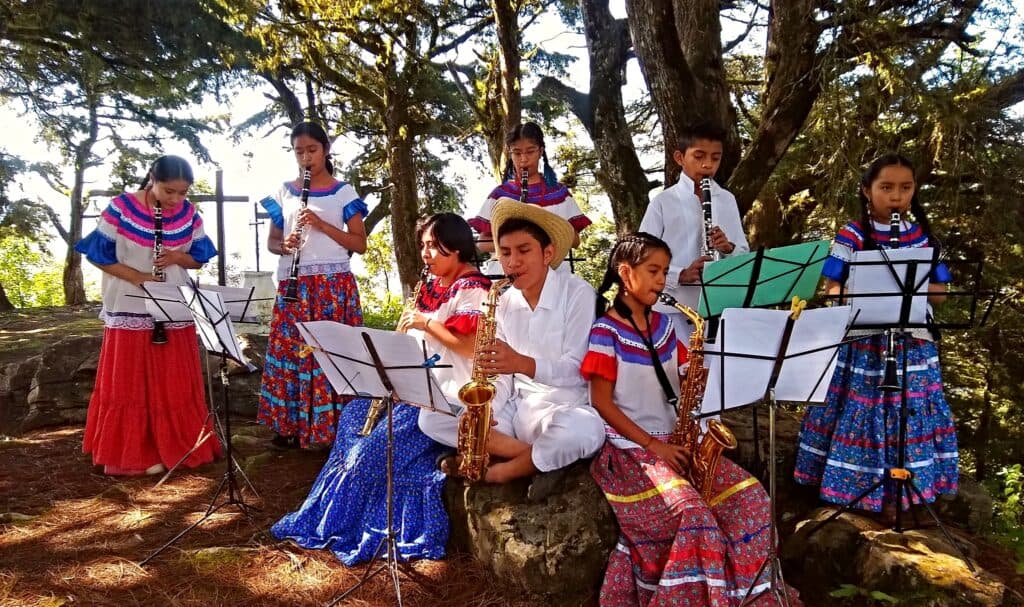 Grupo de jóvenes músicos en trajes tradicionales tocando instrumentos en un entorno natural, promoviendo la educación musical en México.