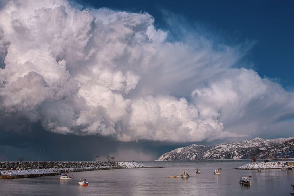 Nubes densas y amenazantes sobre un puerto, con barcos pesqueros y montañas al fondo, reflejando condiciones climáticas inusuales.
