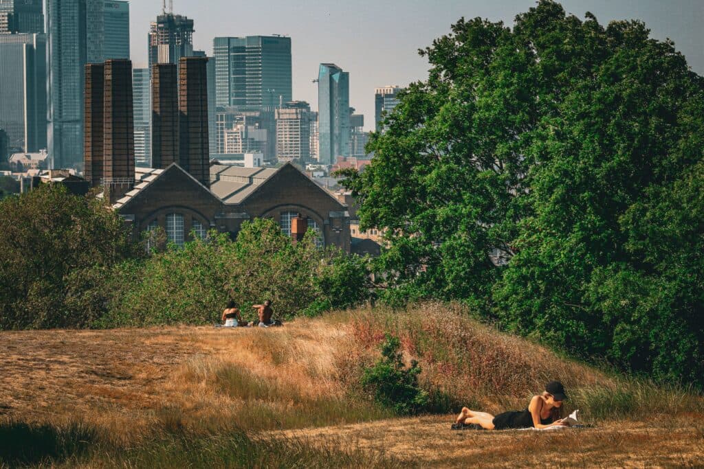 Personas descansando en un parque de Londres bajo el sol, con edificios modernos al fondo, reflejando la ola de calor extremo.