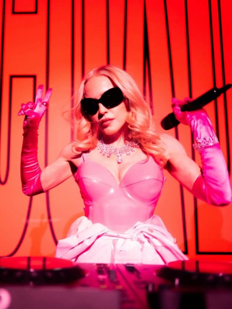 Madonna en un escenario con fondo rojo, luciendo un atuendo rosa y gafas oscuras, sosteniendo un micrófono y mostrando el signo de paz.