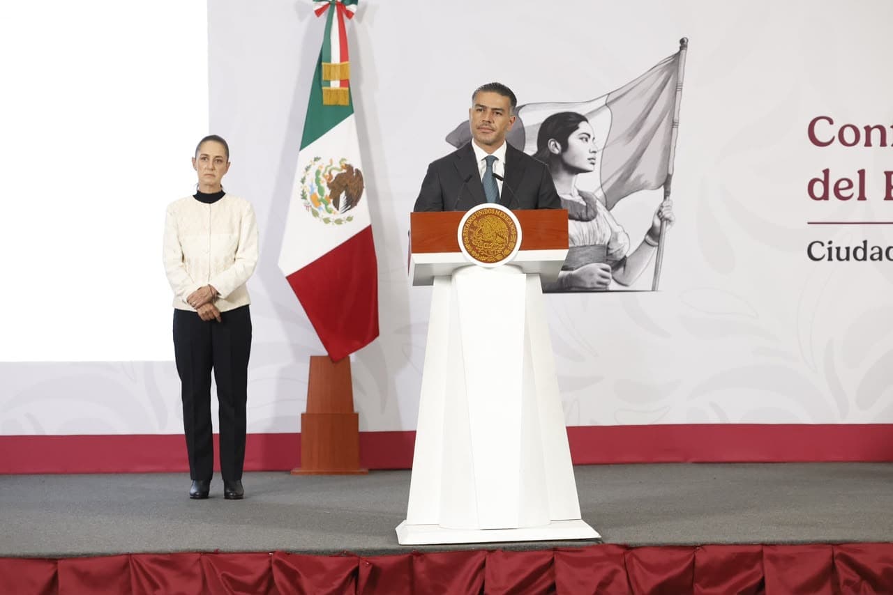 Omar García Harfuch, secretario de Seguridad, habla en conferencia mientras una mujer lo acompaña, con la bandera de México de fondo.