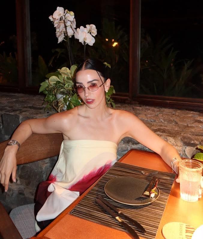 Estíbaliz Badiola posando en un restaurante, con un vestido sin mangas y gafas de sol, rodeada de plantas y decoración elegante.