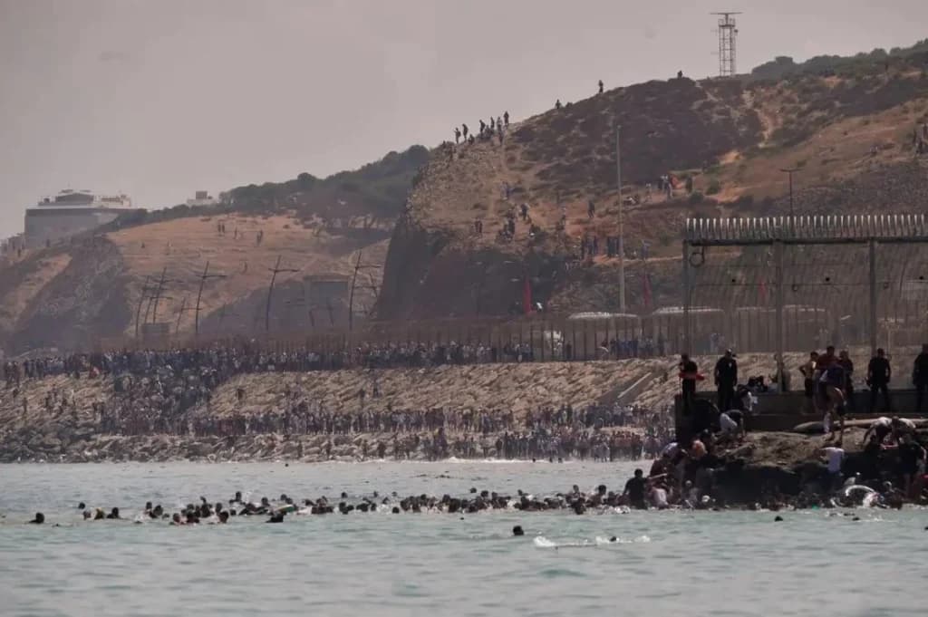 Migrantes en el mar intentando cruzar hacia Ceuta, mientras en la costa se observa una multitud en la frontera con Marruecos.