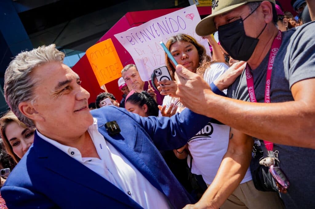 Hombres y mujeres de izquierda en Baja California apoyan a Jesús Alejandro Ruiz Uribe durante un evento, con carteles de bienvenida.