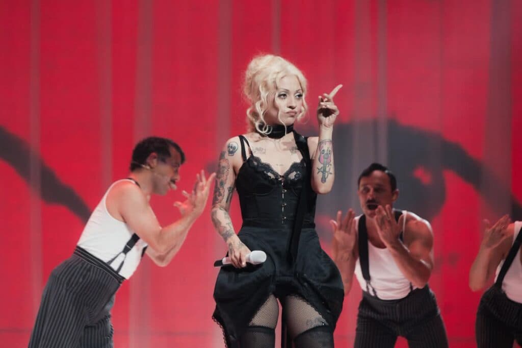 Mon Laferte en el escenario durante una actuación, luciendo un vestido negro y rodeada de bailarines en un ambiente vibrante.