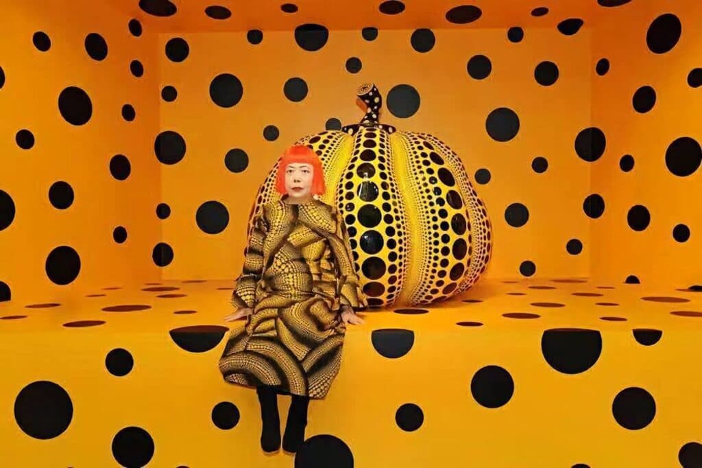 Yayoi Kusama posando en una sala decorada con lunares amarillos y negros, junto a una calabaza gigante, símbolo de su arte contemporáneo.