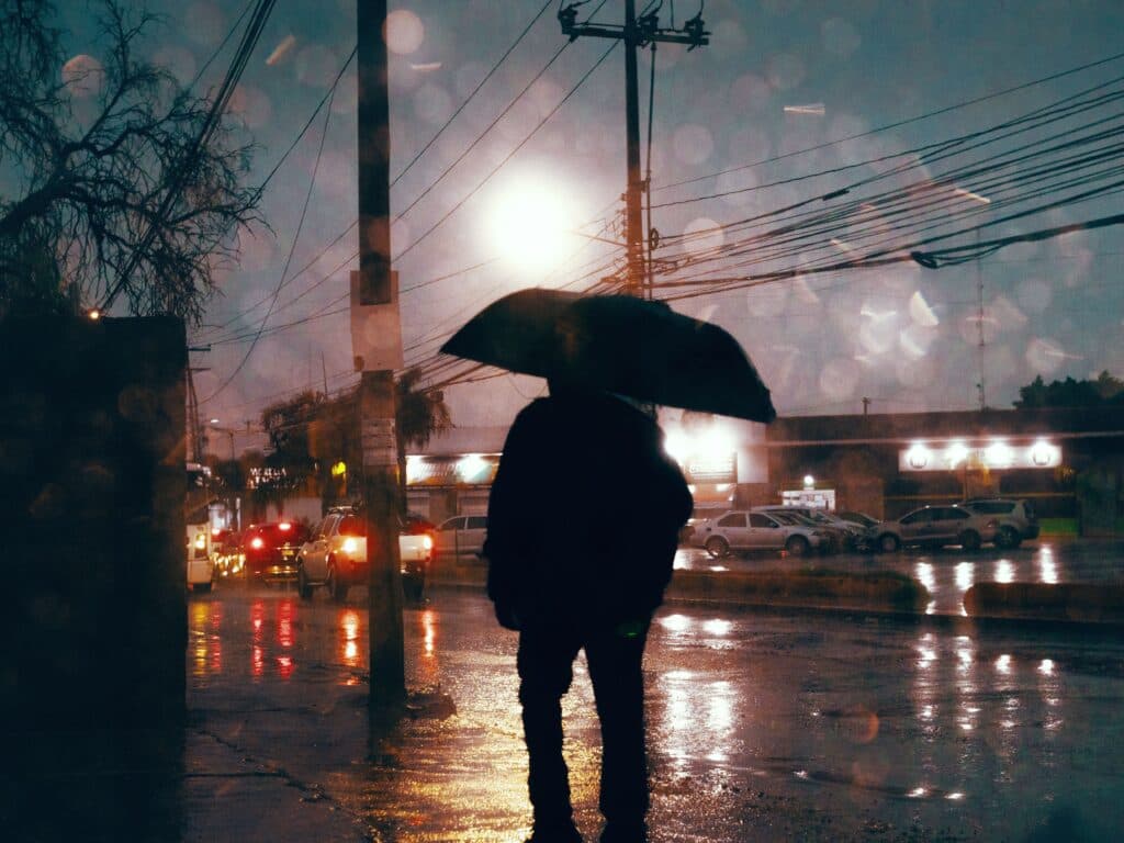 Persona con paraguas en una calle de la CDMX durante una tormenta eléctrica, con lluvia intensa y luces de automóviles al fondo.