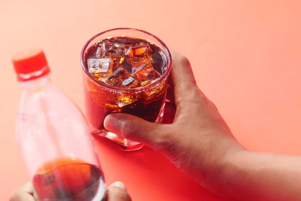 Un vaso con refresco y hielo en una mano, junto a una botella de bebida azucarada, resaltando el riesgo para la salud del hígado.