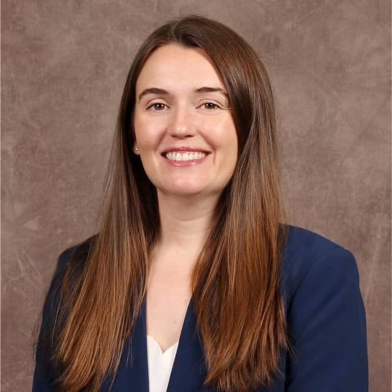 Mujer con cabello largo y lacio, vestida con blazer oscuro, sonríe frente a un fondo neutro, destacando su rol como