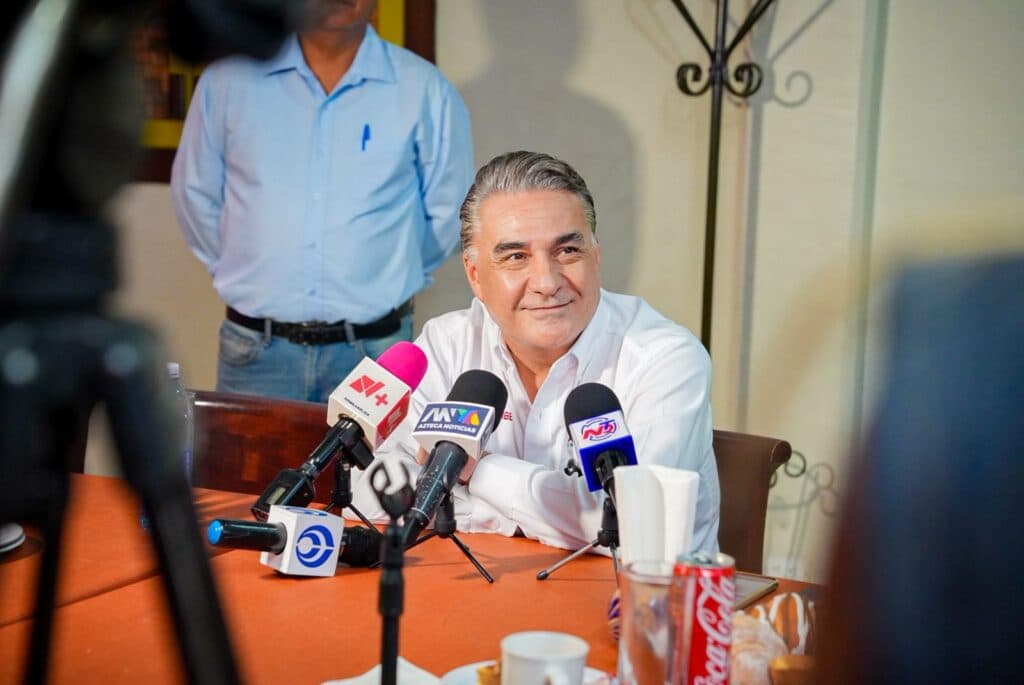 Jesús Ruiz Uribe en conferencia de prensa, rodeado de micrófonos, reafirmando su apoyo a los programas sociales en Baja California.