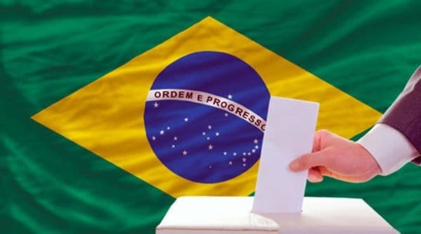Una mano deposita un voto en una urna, con la bandera de Brasil de fondo, en el contexto de las elecciones presidenciales.