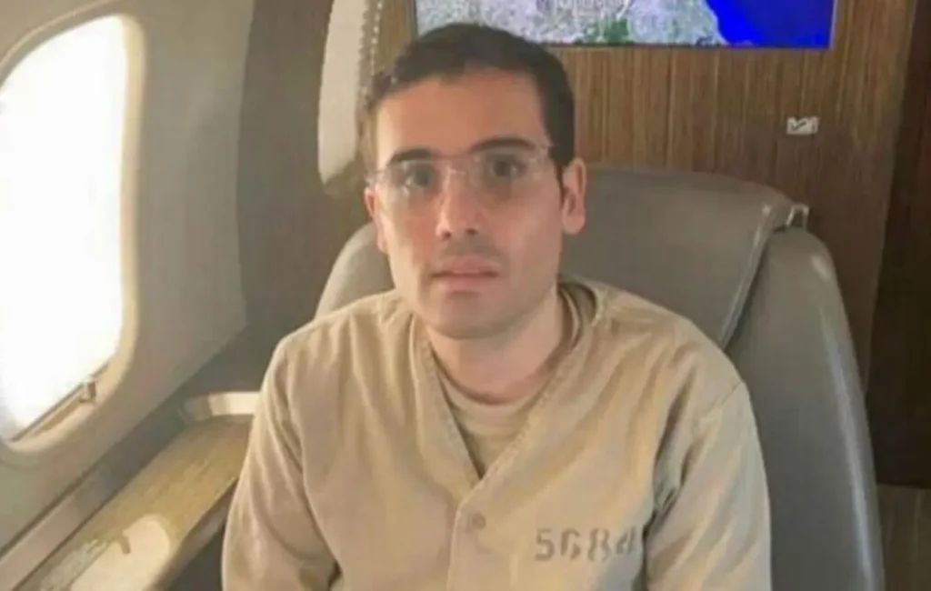 Ovidio Guzmán, hijo de Joaquín "El Chapo" Guzmán, sentado en un avión, vestido con uniforme de prisión, en contexto de su audiencia en EE. UU.